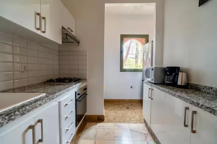 Kitchen of a vintage apartment in Marrakech featuring white cabinets, granite countertops, appliances, and a view of trees through the window.