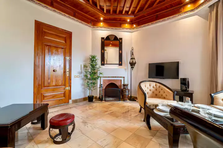 Warm, traditional living room of a vintage apartment in Marrakech, featuring a carved wooden ceiling, fireplace, retro armchairs, and marble flooring.