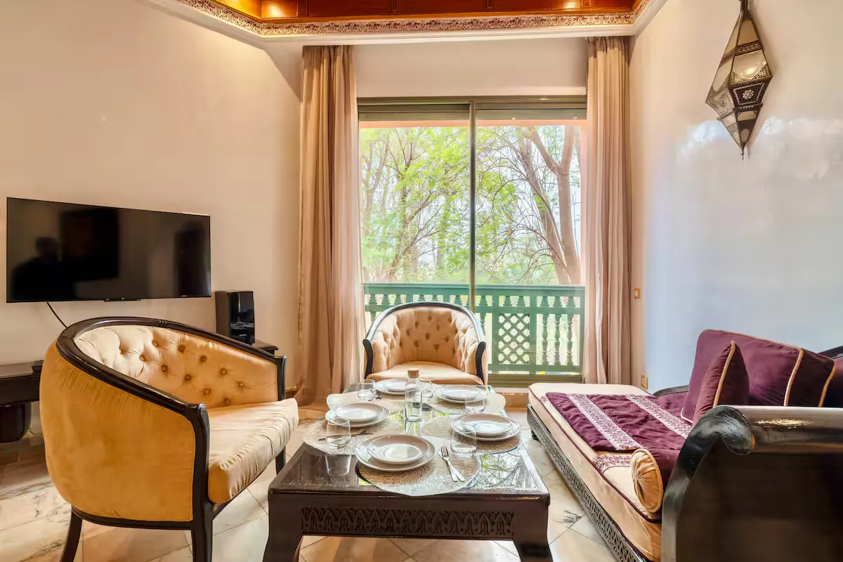 Dining area and living room of a vintage apartment in Marrakech, featuring tufted armchairs, a traditional-style sofa, a dark wood coffee table, and balcony access.
